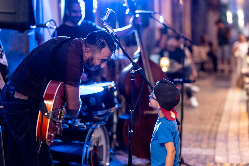 Musicien se penchant vers un enfant devant un micro vintage lors d’un concert en terrasse chez Gourmet & Glouton à Chaudes-Aigues, illustrant un moment de partage et de transmission.